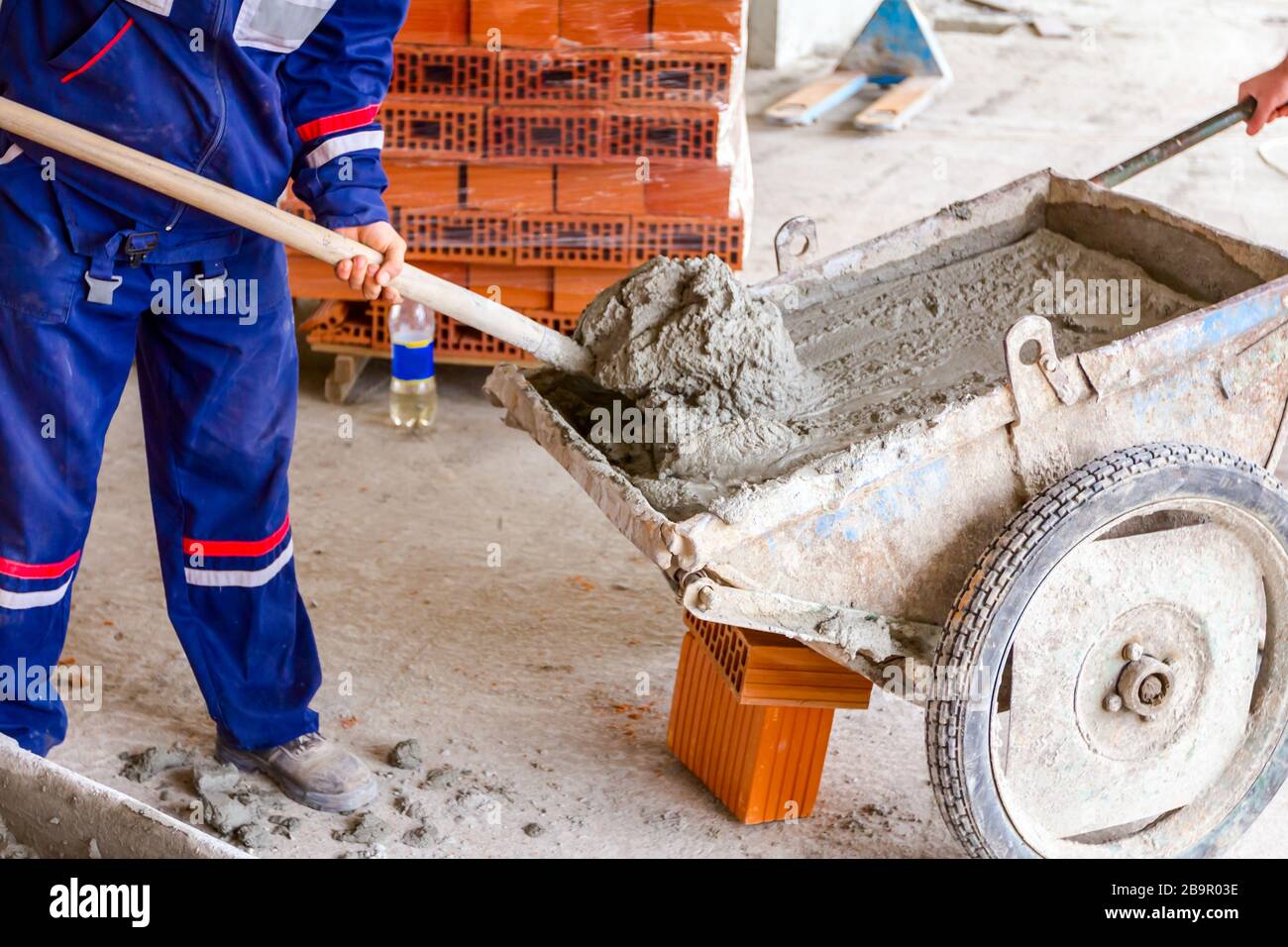 Construction worker is using shovel to unloading cement, mortar, from wheelbarrow on construction site. Stock Photo