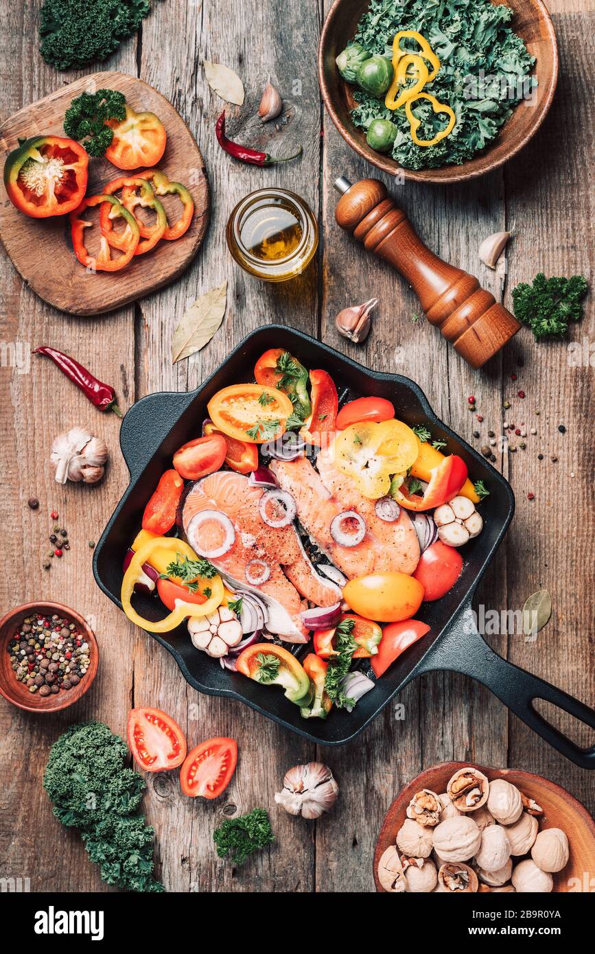 Salmon steak with vegetables on iron grill pan, bowl with kale salad, nuts, organic vegetable and kitchen utensils over wooden background. Top view. R Stock Photo