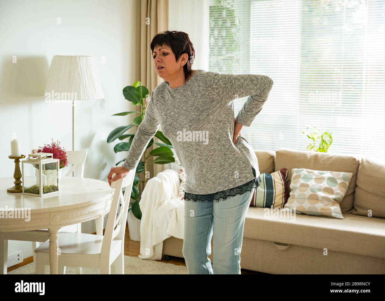 Mature woman suffering from backache at home. Massaging lower back with hand, feeling exhausted, standing in living room. Stock Photo