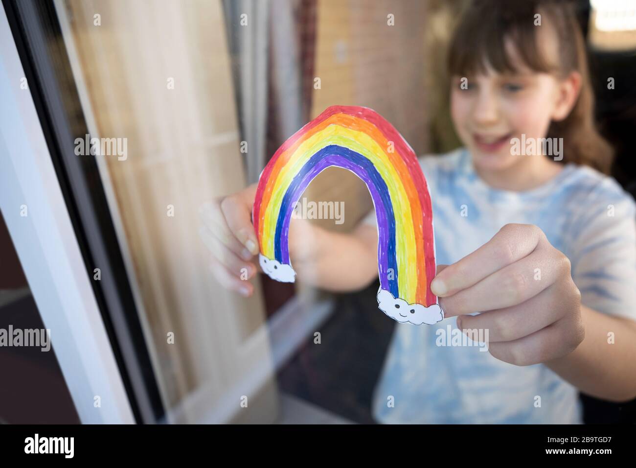 Girl Putting Picture Of Rainbow In Window At Home During Coronavirus Pandemic To Entertain Children Stock Photo