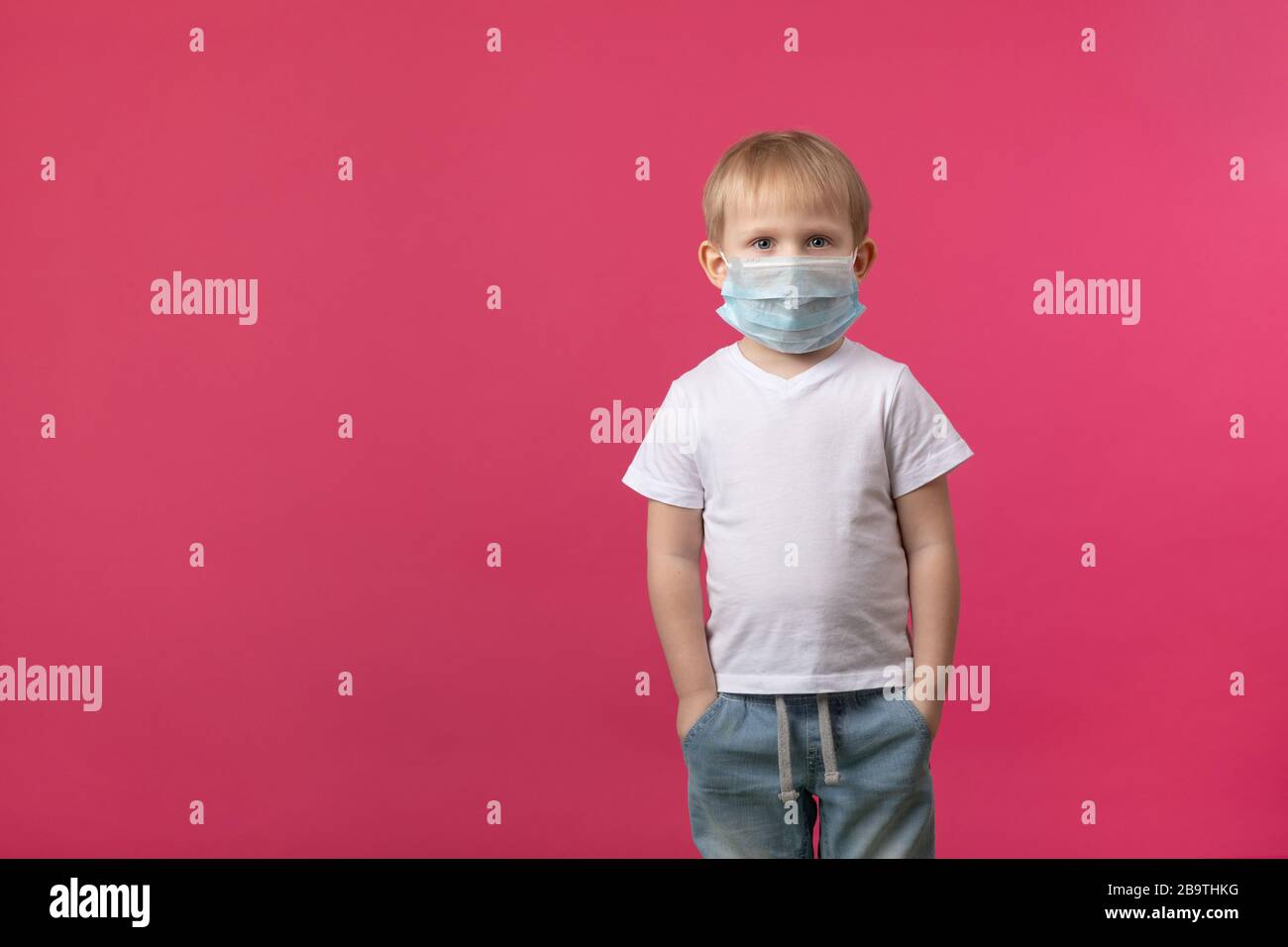 A blond European-looking child boy stands in a medical mask from a coronavirus and looks at the camera. Studio shot on a pink background with place fo Stock Photo