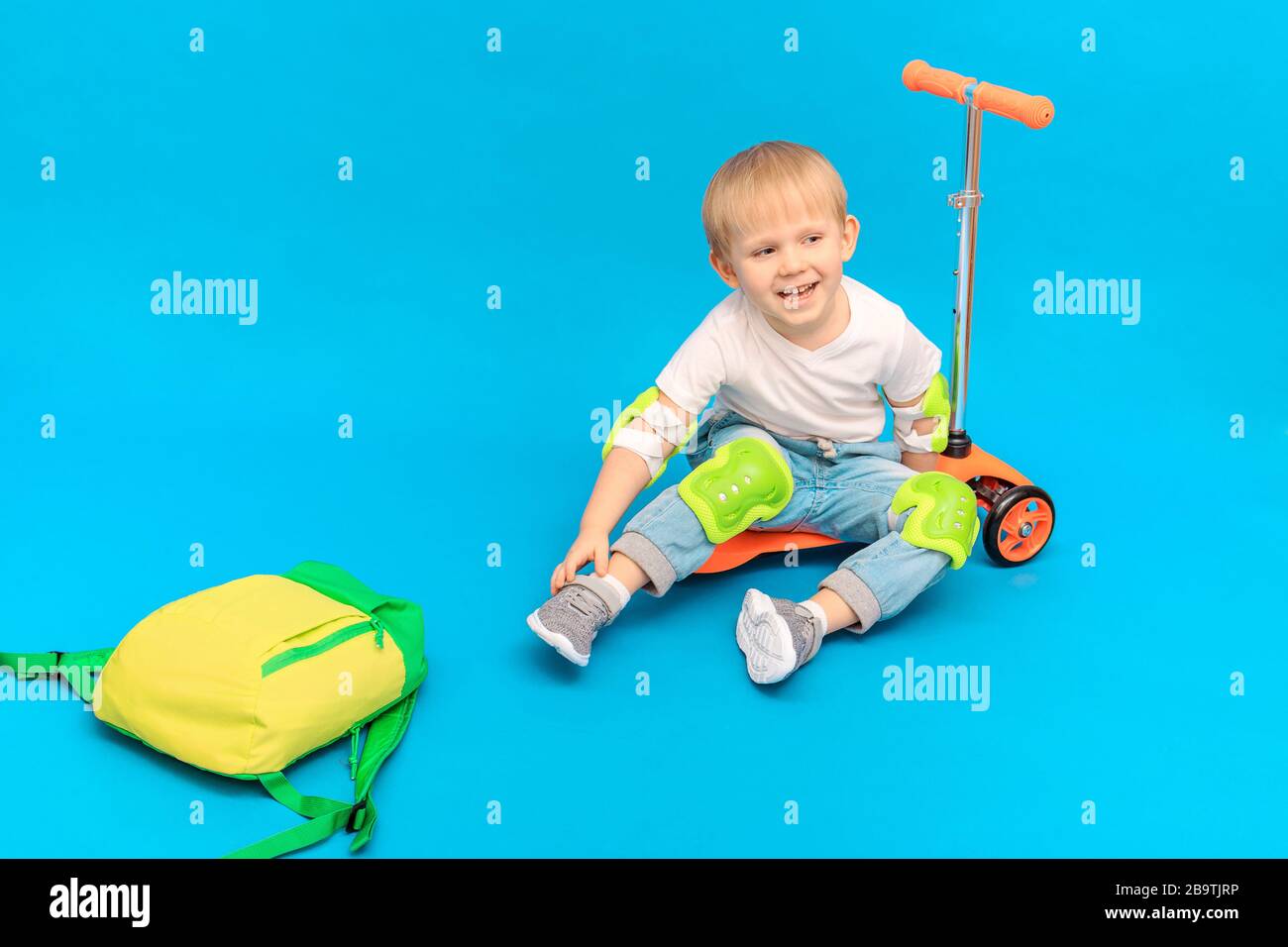 A child boy sits on a scooter in protection against abrasions and bruises, knee pads and elbow pieces and laughs. Studio photography with place for te Stock Photo