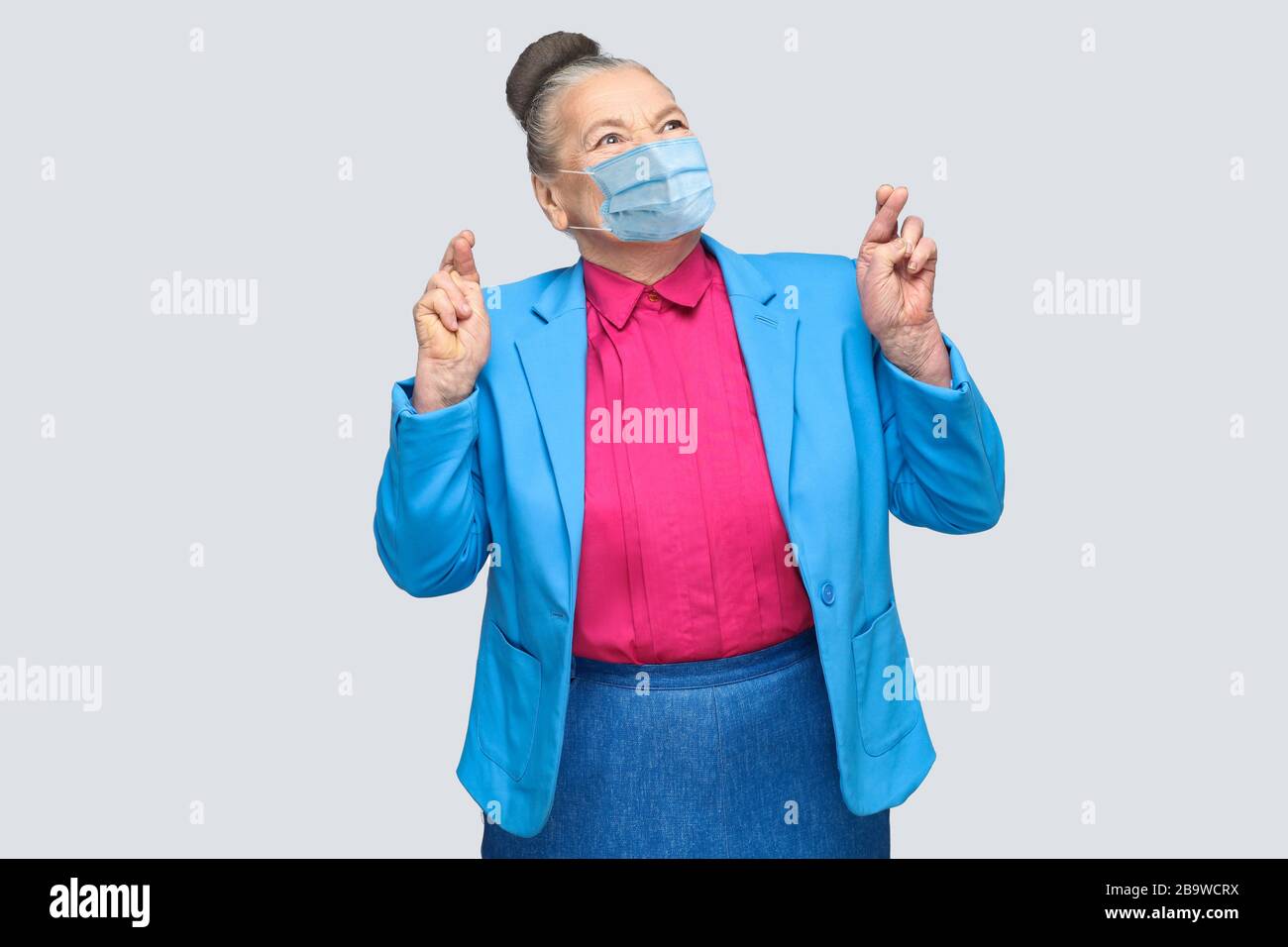 Hopeful with surgical medical mask woman crossed fingers and wish. Grandmother with light blue suit and pink shirt standing with collected bun hair. i Stock Photo