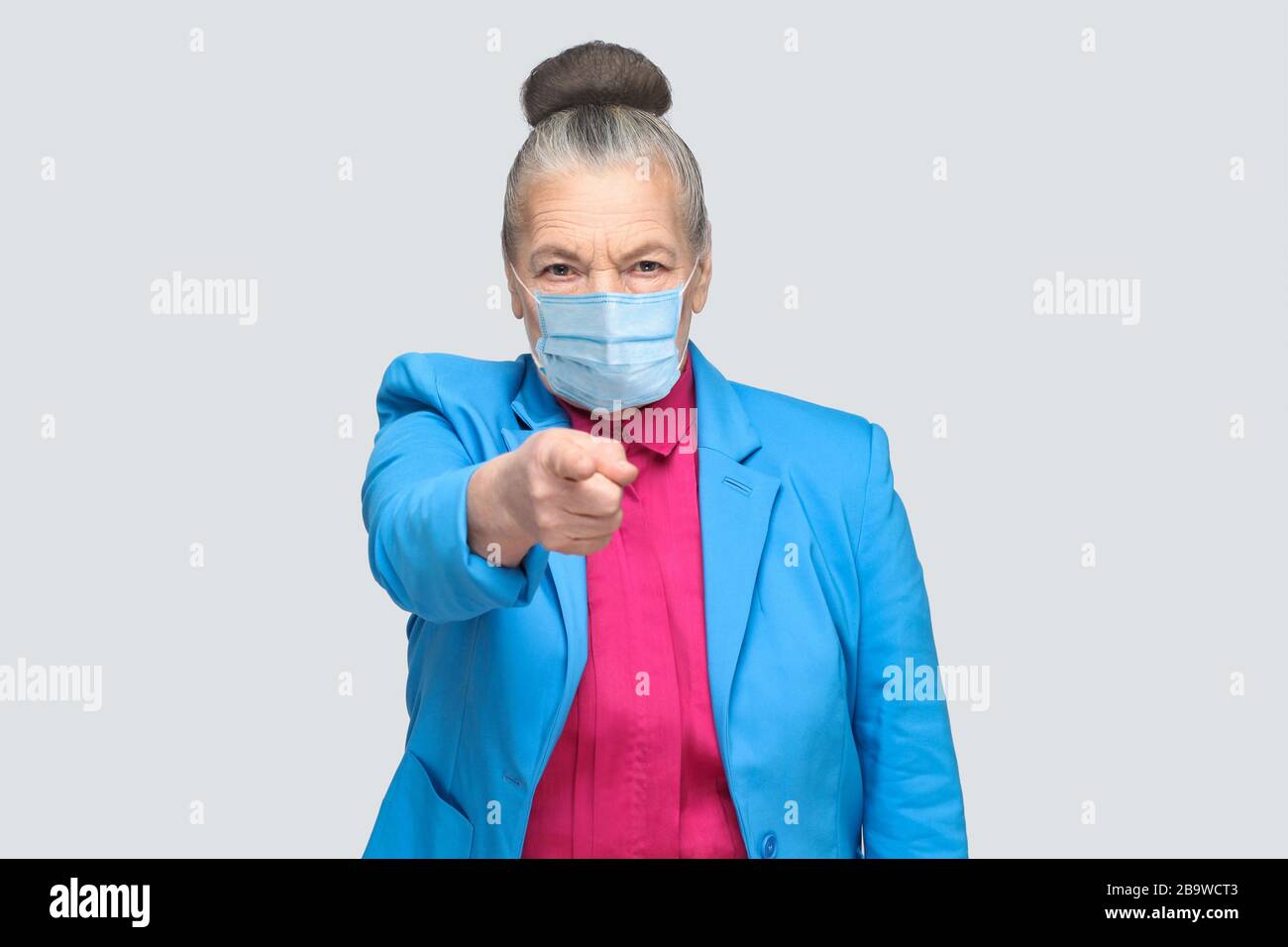Hey you! Woman with surgical medical mask pointing at camera finger with serious face. Portrait of expressive grandmother standing with collected hair Stock Photo