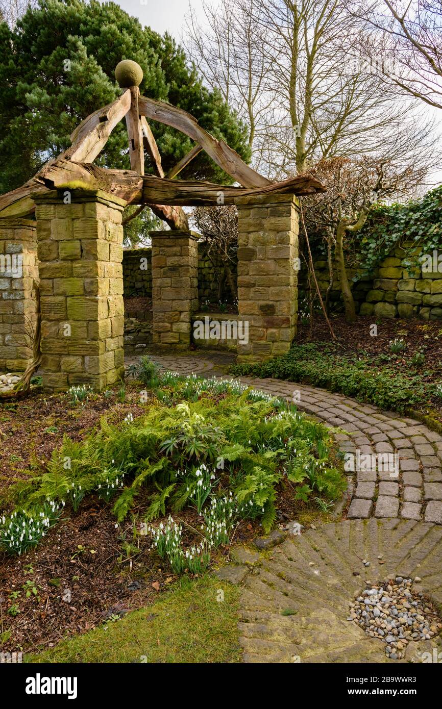Corner of country cottage garden (rustic arbour, timber beams, stone setts, curving path, millstone, snowdrops on border) - York Gate Garden, Leeds UK Stock Photo