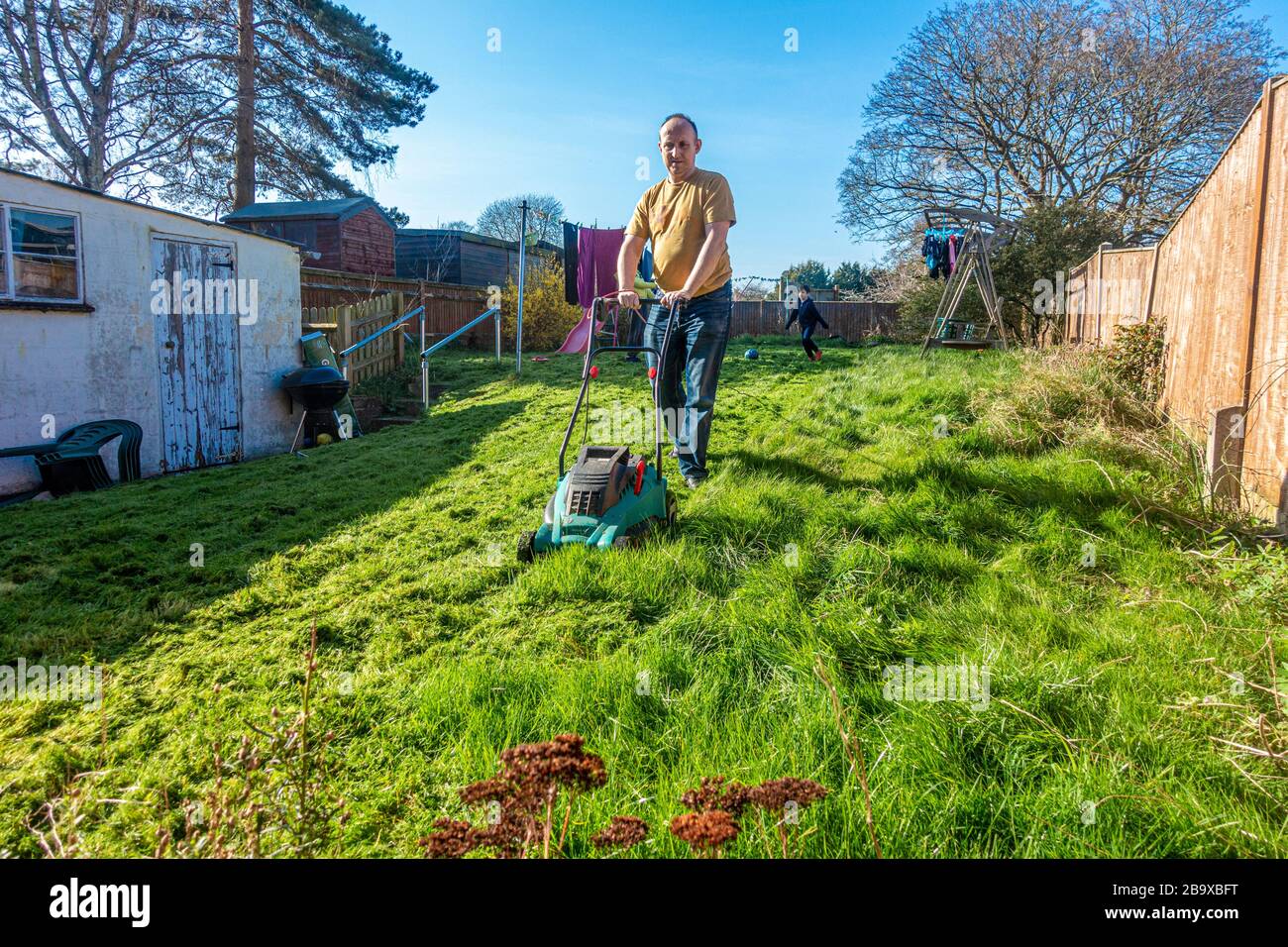 A man cuts the lawn in his back garden for the first time this year. The grass is long and untidy. The man pushes an electric lawnmower Stock Photo
