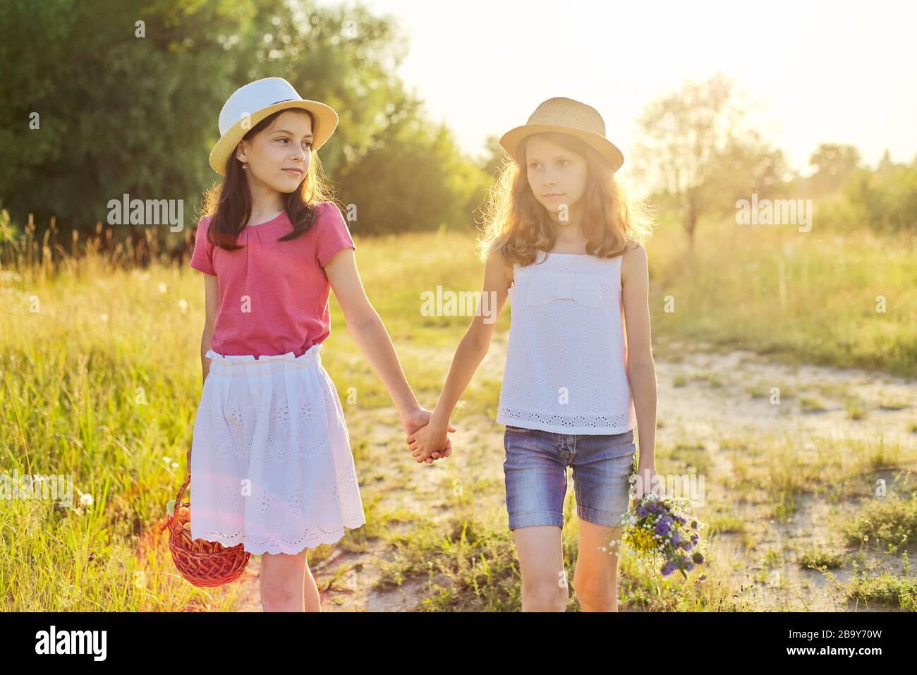 Two girls walking in summer meadow holding hands, children with basket of cherries bouquet of wildflowers, with sunset Stock Photo