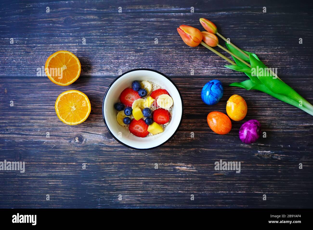 Breakfast cereal bowl with fresh fruits (strawberry, banana, blueberry, mango). Half-cut orange, colorful Easter eggs and orange tulips for decoration Stock Photo