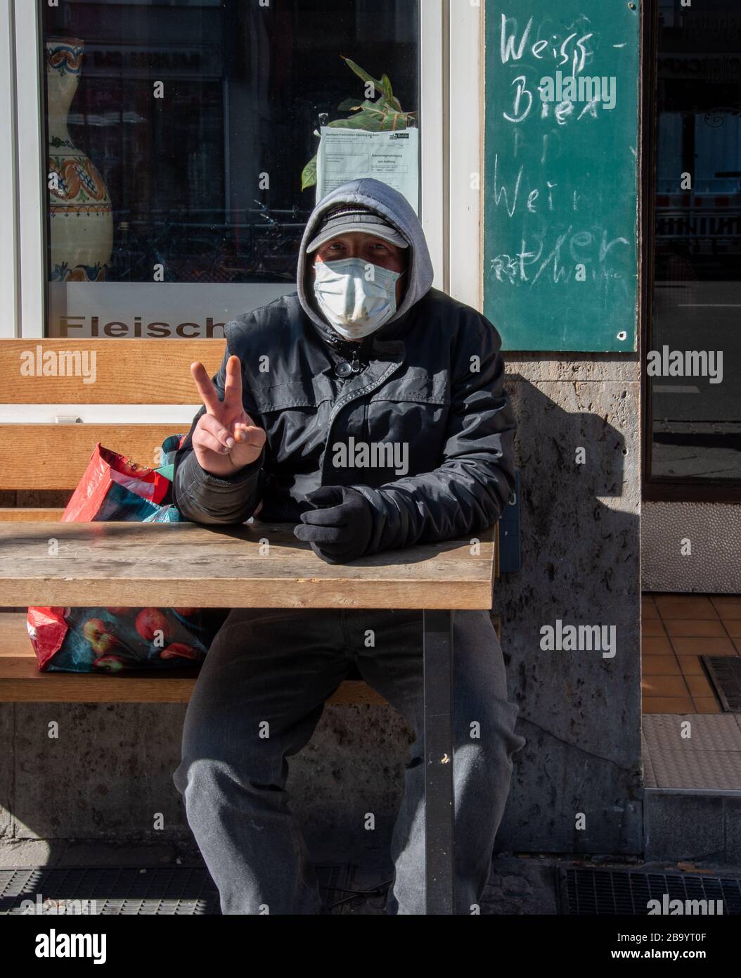 Man wearing a facemark doing a V for Victory sign while resting on a bench outside a closed restaurant in central Berlin during the corona crisis Stock Photo