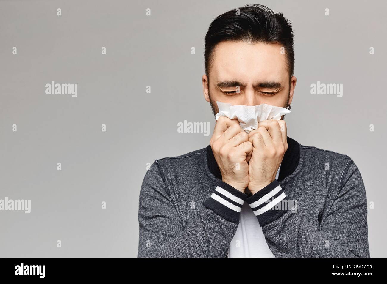 Young man sneezing and covering mouth with paper napkin posing on grey background, isolated. Studio picture of a young man with a handkerchief. Ill Stock Photo