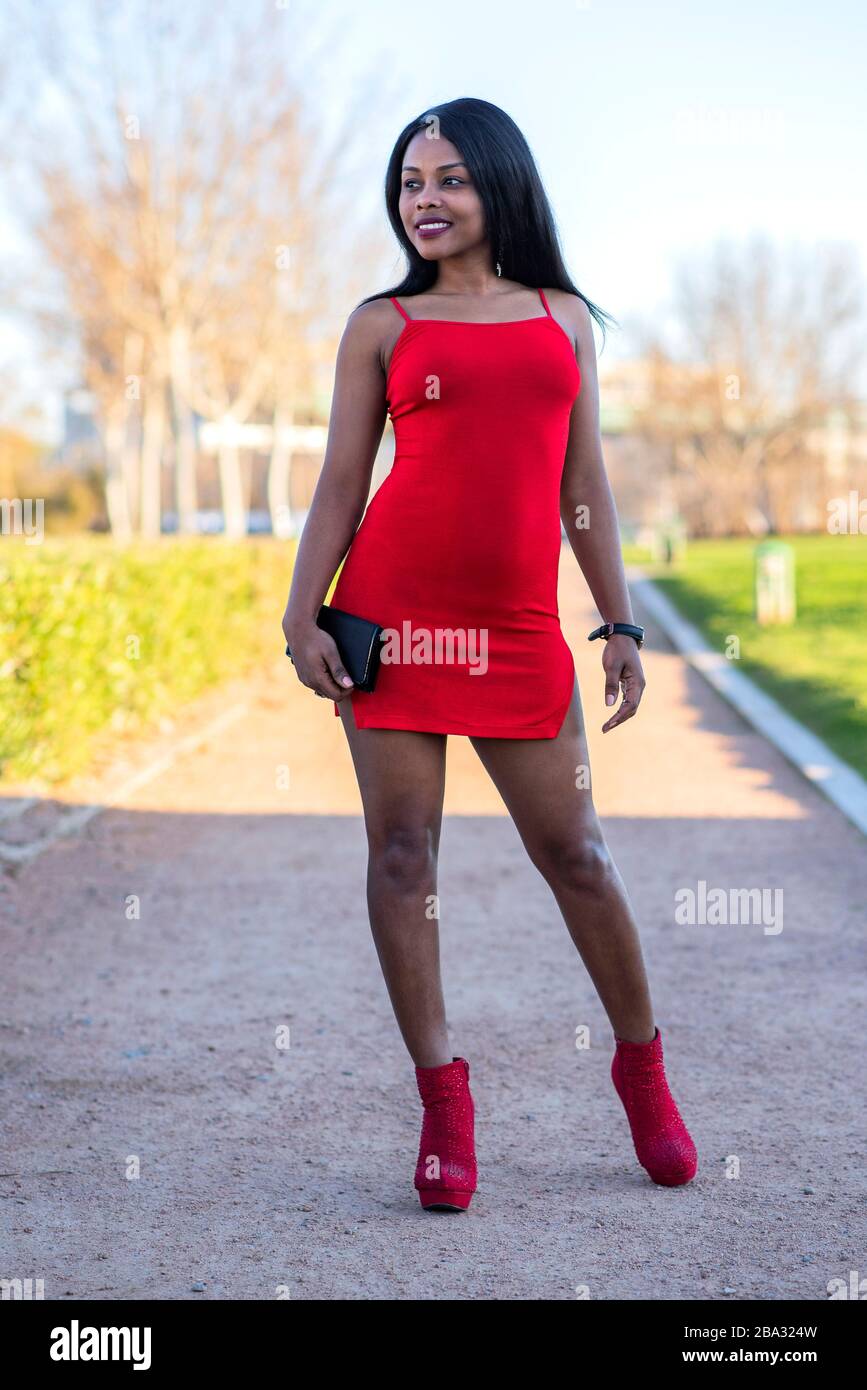 dark-skinned, black-haired woman posing in a red dress and shoes in an outdoor park Stock Photo