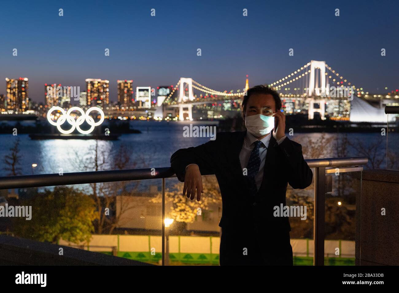 Odaiba, Minato Ward, Tokyo, Japan—MAR 25, 2020: Japanese Businessman with a Mask Phoning near the Olympic Symbol during the Corona Virus Pandemic Stock Photo