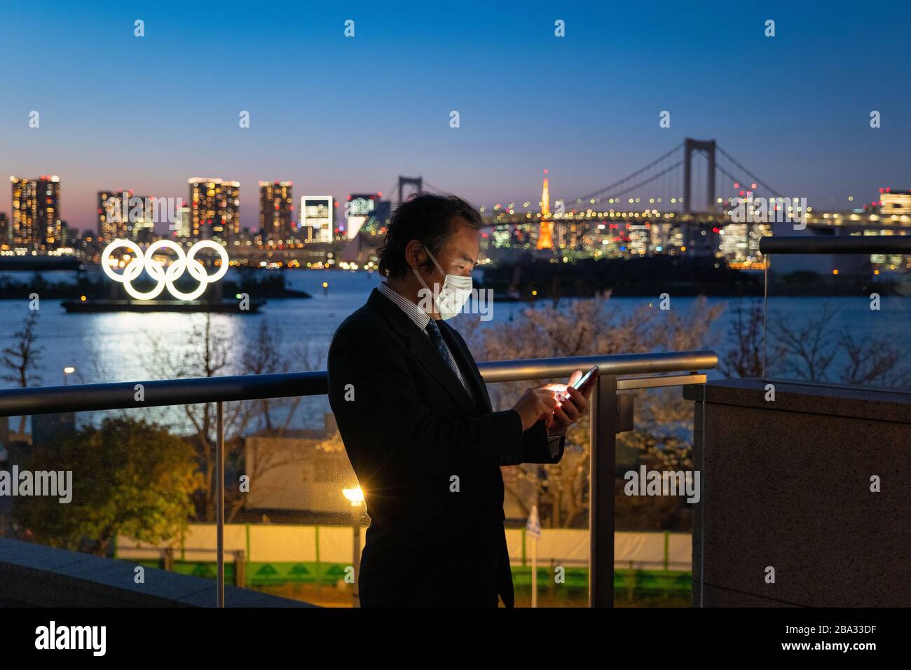 Odaiba, Minato Ward, Tokyo, Japan—MAR 25, 2020: Japanese Businessman with a Mask Texting near the Olympic Symbol during the Corona Virus Pandemic Stock Photo