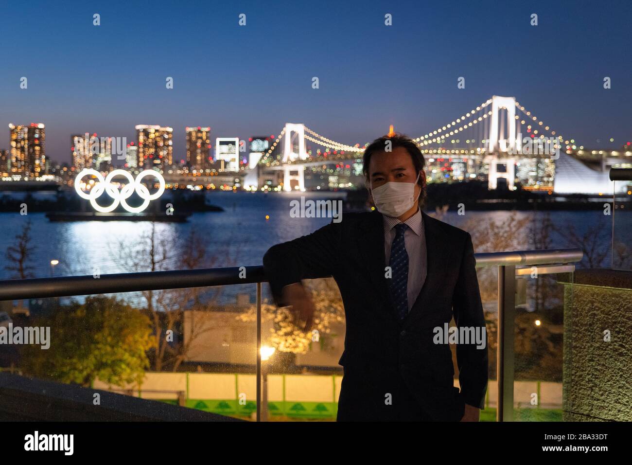 Odaiba, Minato Ward, Tokyo, Japan—MAR 25, 2020: Japanese Businessman Wears a Mask in front of the Olympic Symbol during the Corona Virus Pandemic Stock Photo