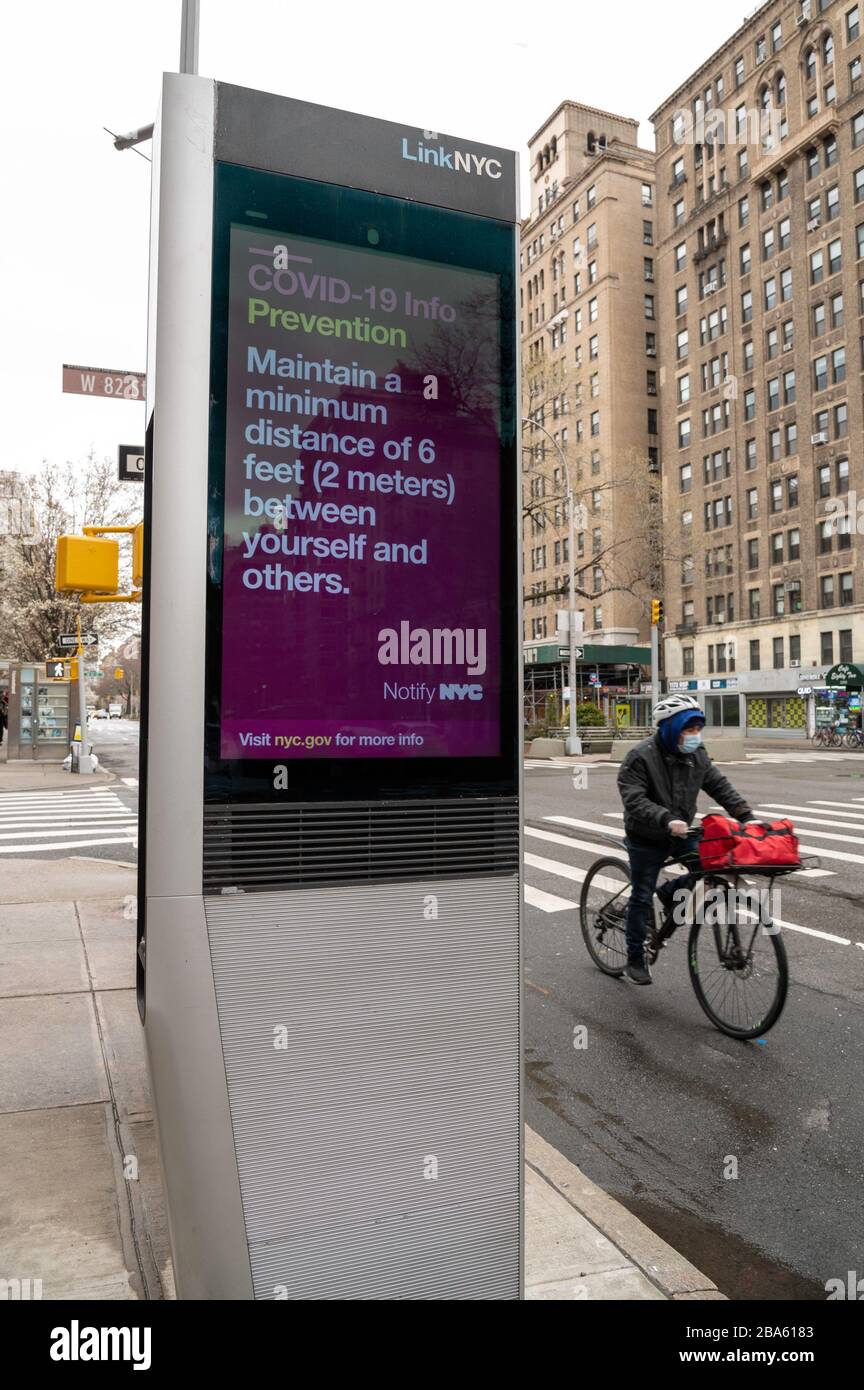 NEW YORK, NY - MARCH 25, 2020. A food delivery worker wearing a protective mask rides his bike past a LinkNYC kiosk on the Upper West Side of Manhattan in New York City. The kiosk displays a public service message regarding the Coronavirus outbreak. The World Health Organization declared coronavirus (COVID-19) a global pandemic on March 11th. Stock Photo