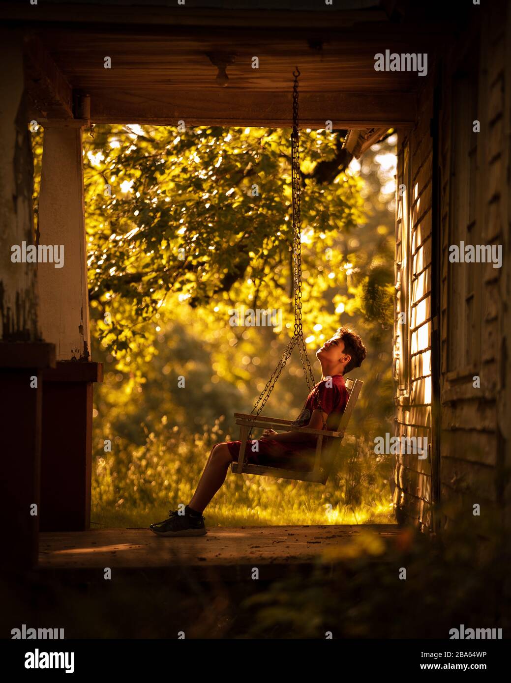 A young boy sits on a country porch swing exploring an abandoned farm house in the Ozark Mountains. Stock Photo