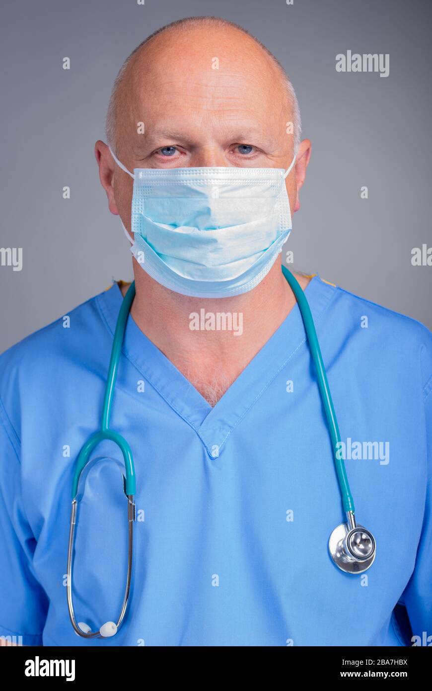 Portrait of a caucasian hospital doctor in blue scrubs, looking at the viewer & wearing a surgical mask with a stethoscope, against a grey background. Stock Photo