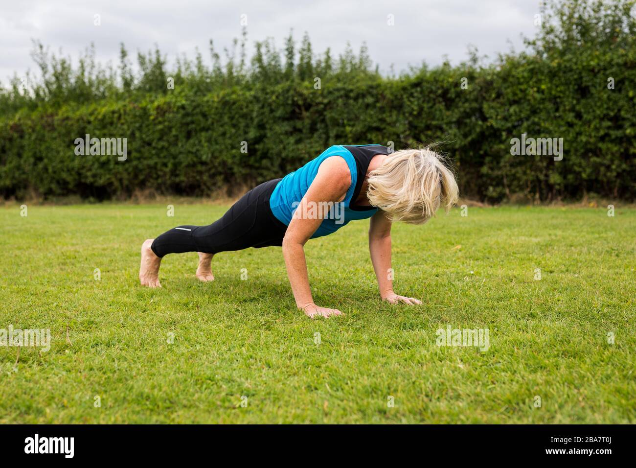 A middle aged woman practicing yoga barefoot outside in a grassy park. She is wearing a bright blue vest and black leggings. The style of yoga she is Stock Photo