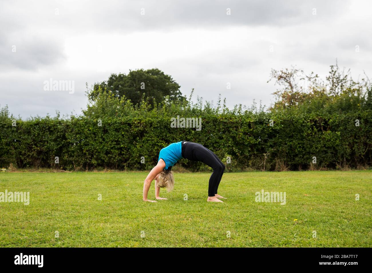 A middle aged woman practicing yoga barefoot outside in a grassy park. She is wearing a bright blue vest and black leggings. The style of yoga she is Stock Photo