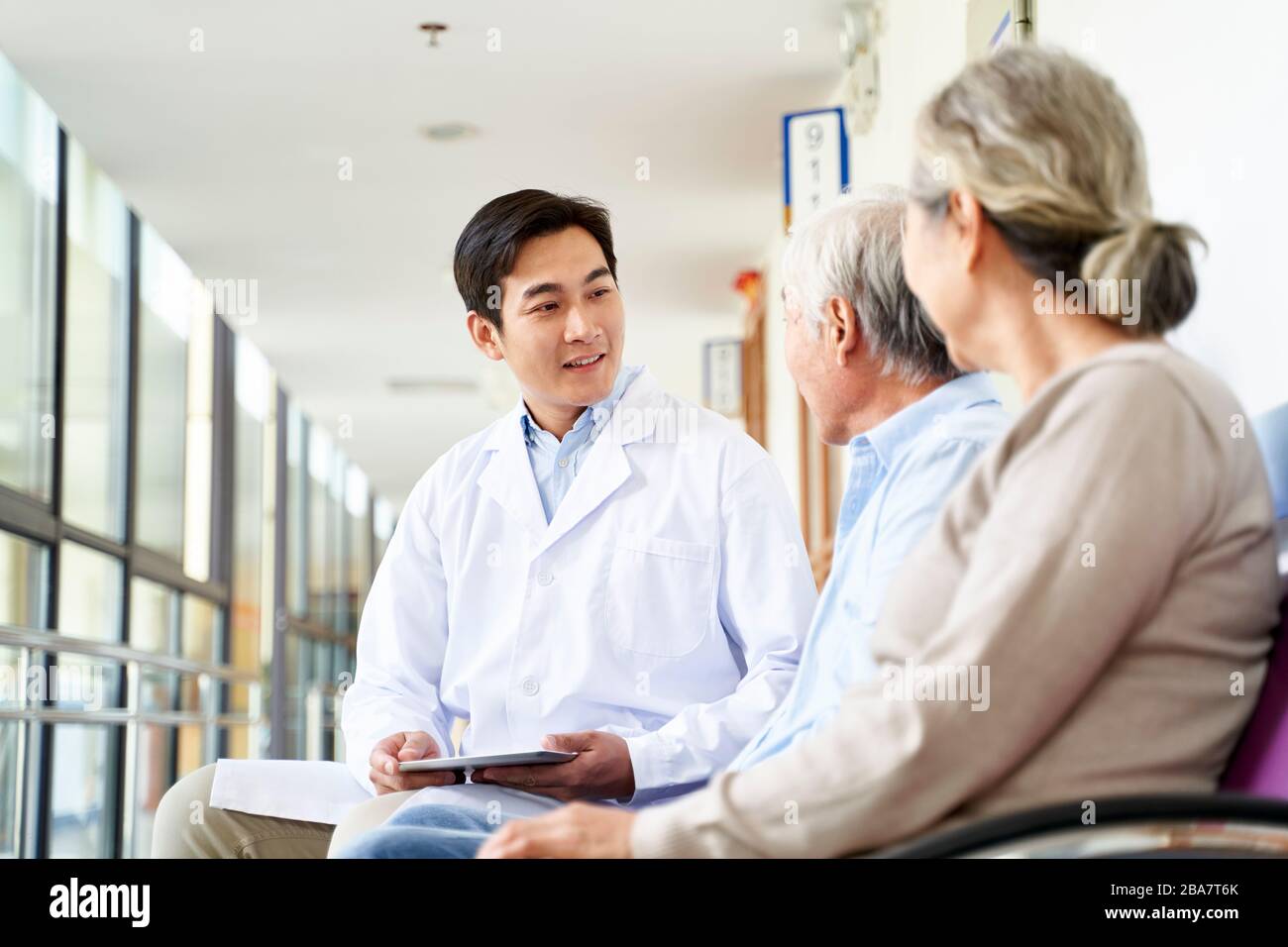 friendly young asian doctor talking to old couple in hospital hallway Stock Photo
