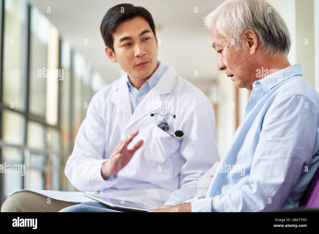 young asian doctor talking and explaining test result and diagnosis to demoralized elderly patient in hospital hallway Stock Photo