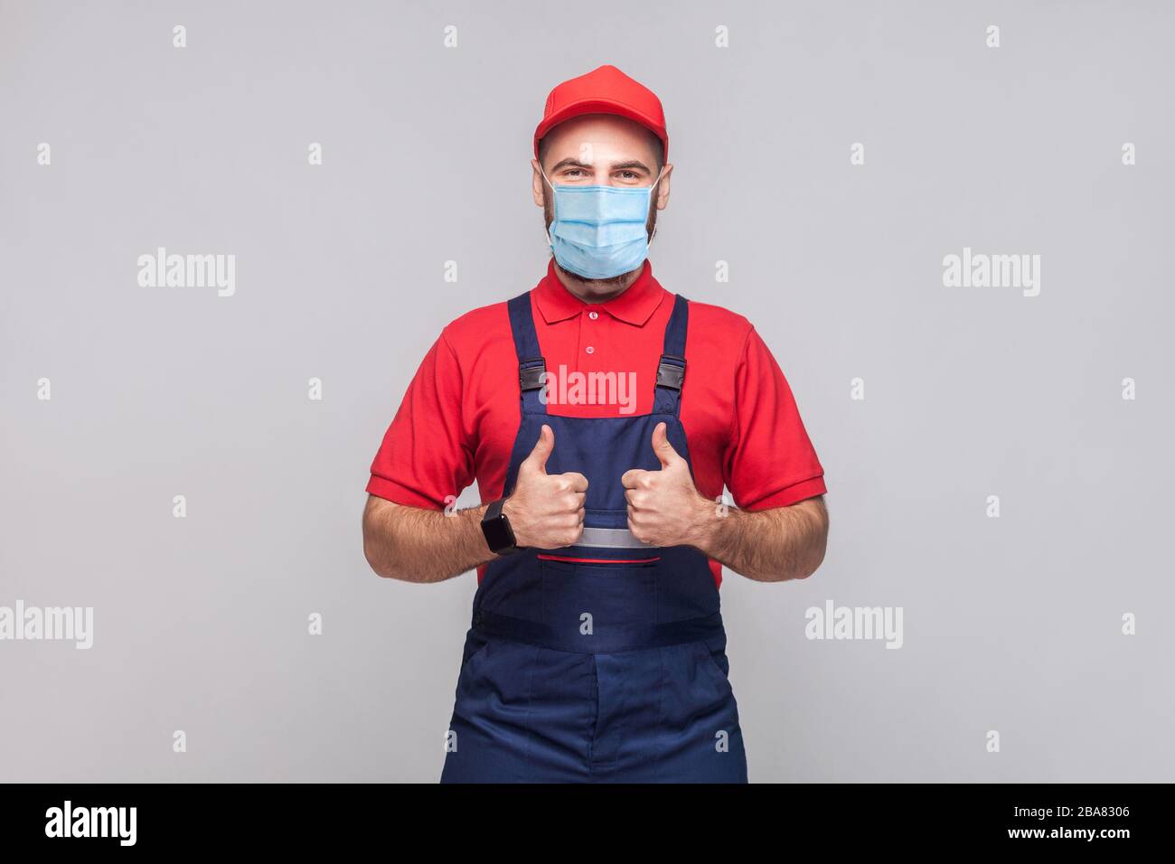 Work are done! Portrait of young man with surgical medical mask in blue overall, red t-shirt , cap, standing and showing thumps up and looking at came Stock Photo