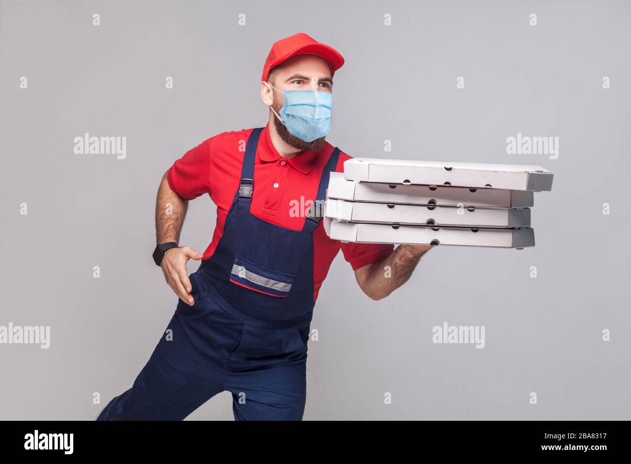 Delivery pizza on quarantine. Hurry up! Young man with surgical medical mask in blue uniform and red t-shirt holding stack pizza boxes and running to Stock Photo