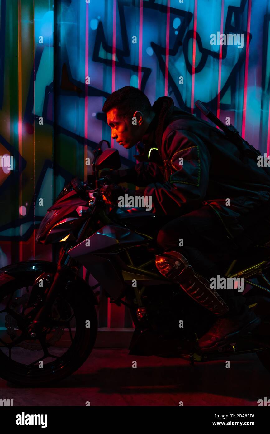 side view of mixed race cyberpunk player riding motorcycle on street with graffiti Stock Photo