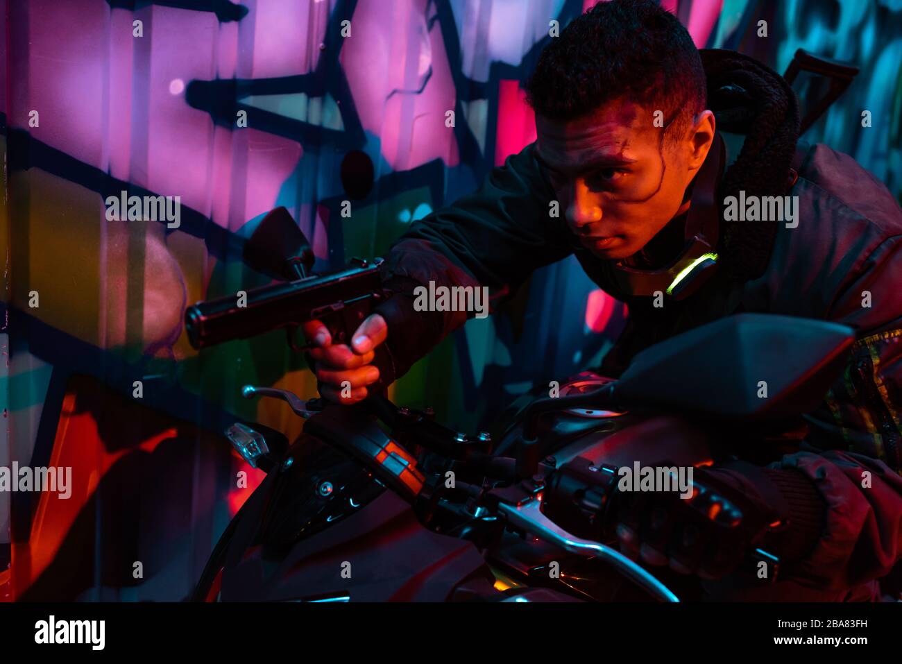 mixed race cyberpunk player on motorcycle aiming gun on street with graffiti Stock Photo