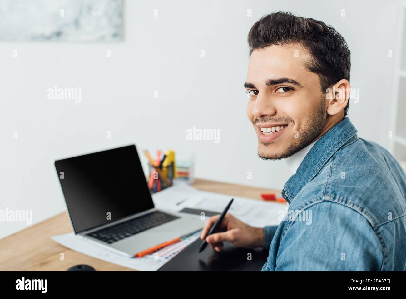 Smiling ux designer looking at camera while using graphics tablet and laptop at table Stock Photo