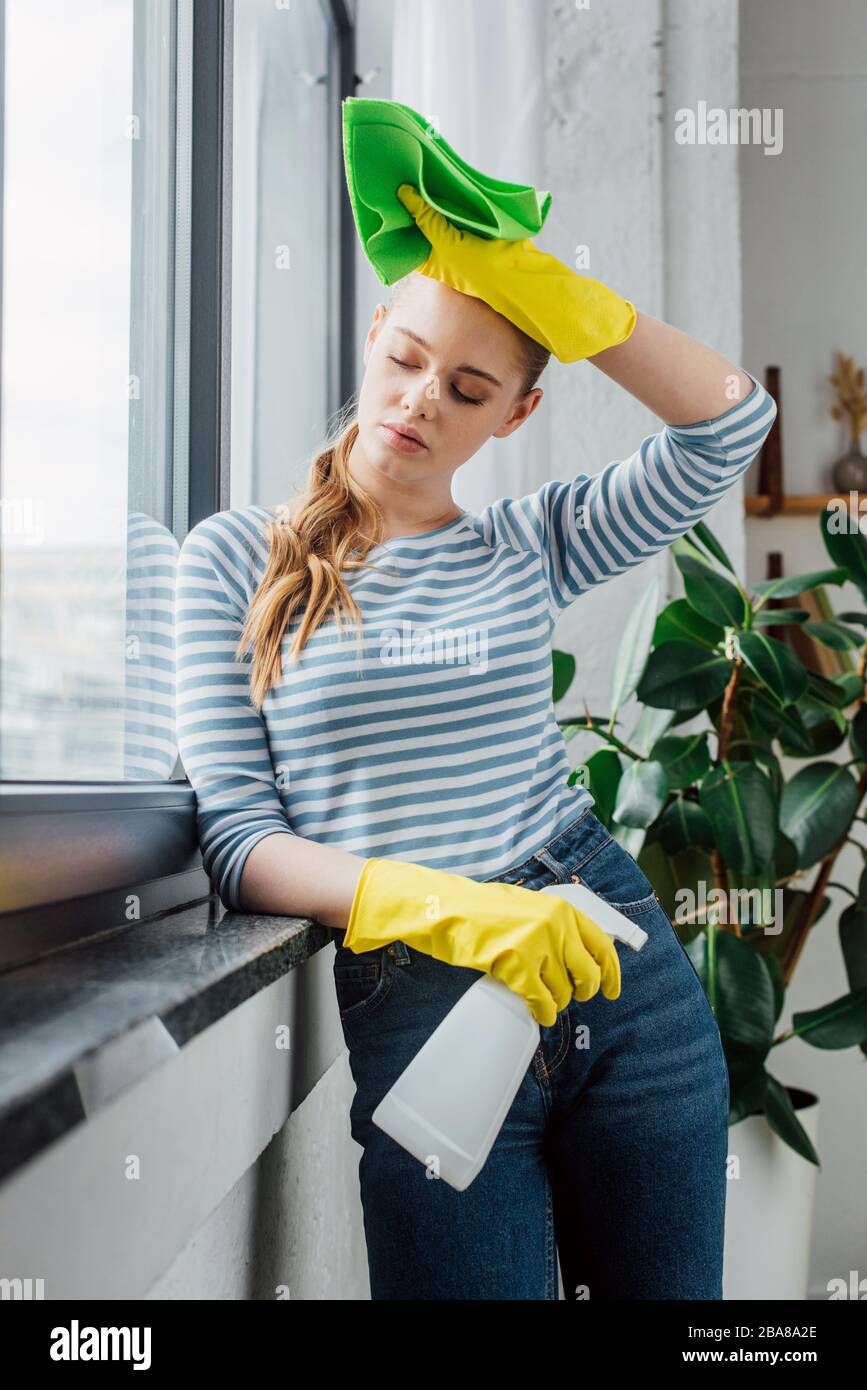 Tired girl in rubber gloves holding detergent and rag near window at ...
