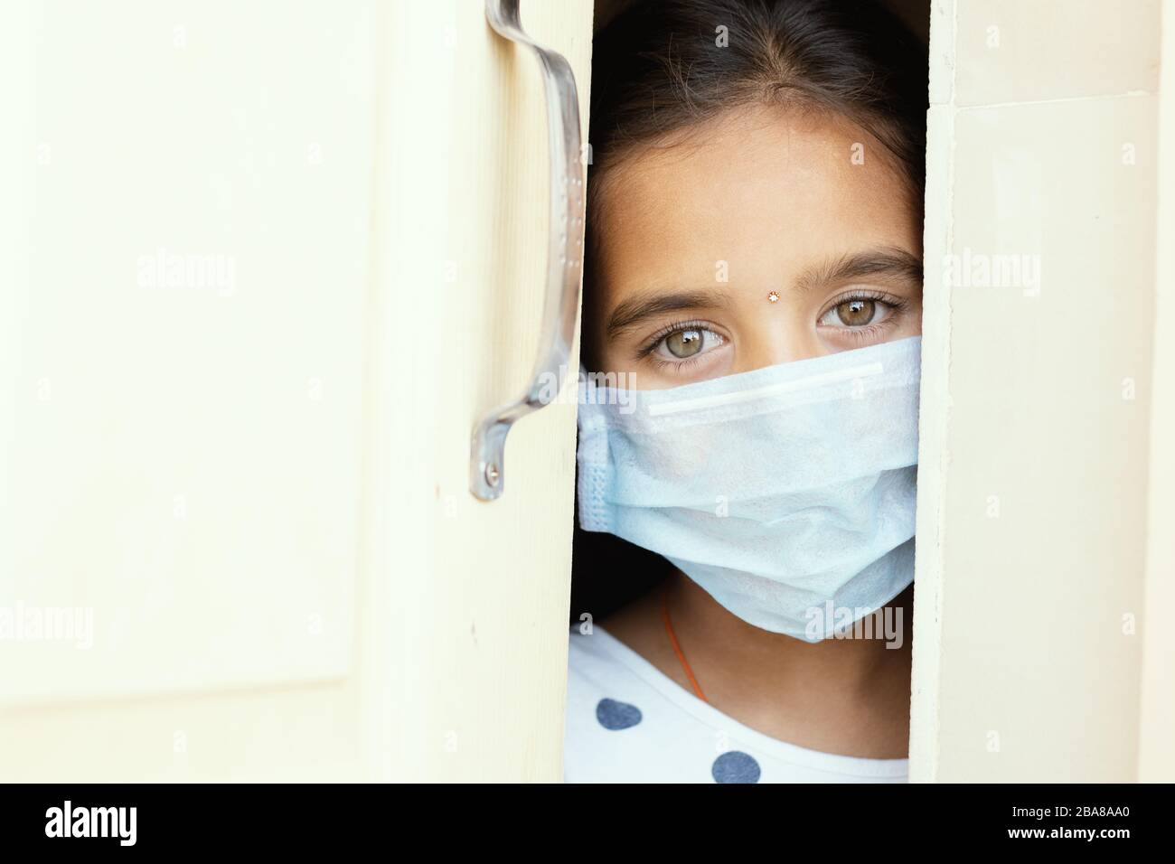 Young sad girl with medical mask wearing sneaking out through the home door - concept of home quarantine due to covid-19 or coronavirus outbreak. Stock Photo