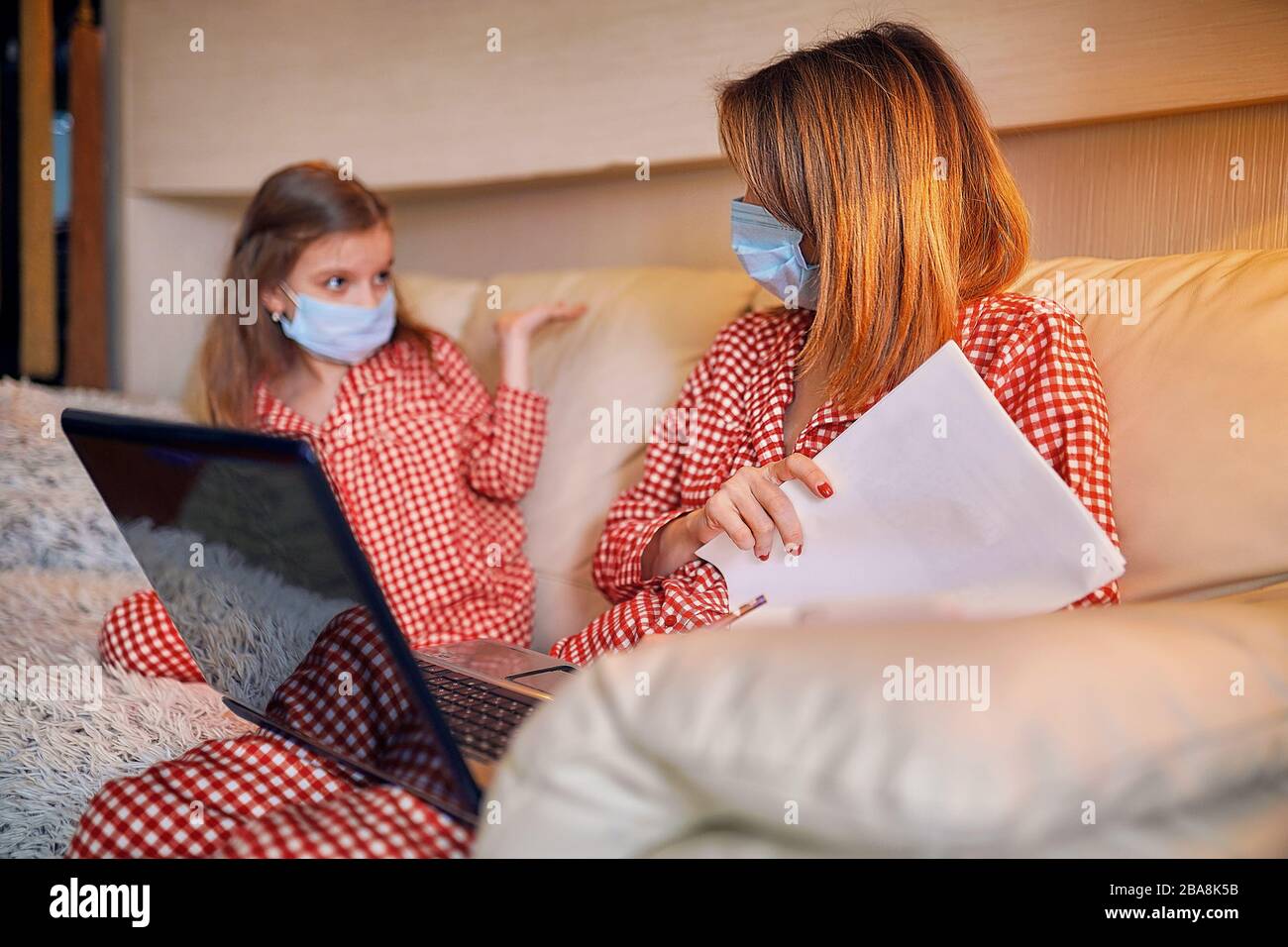 Woman in pajamas with notebook and papers working from home wearing protective mask while her kid, daughter playing computer console games. Stock Photo