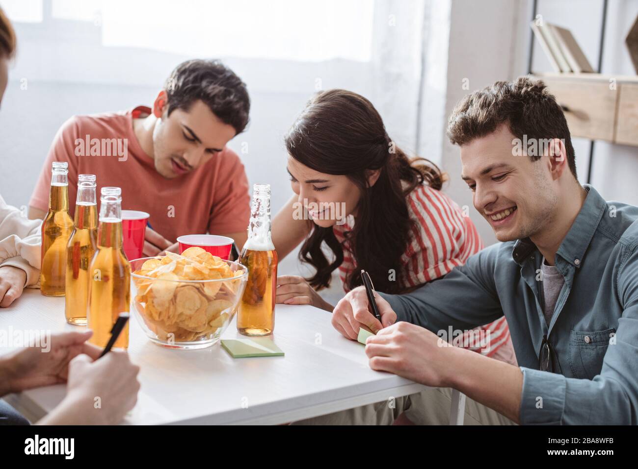 cheerful friends writing on sticky notes while playing name game at table with drinks and chips Stock Photo
