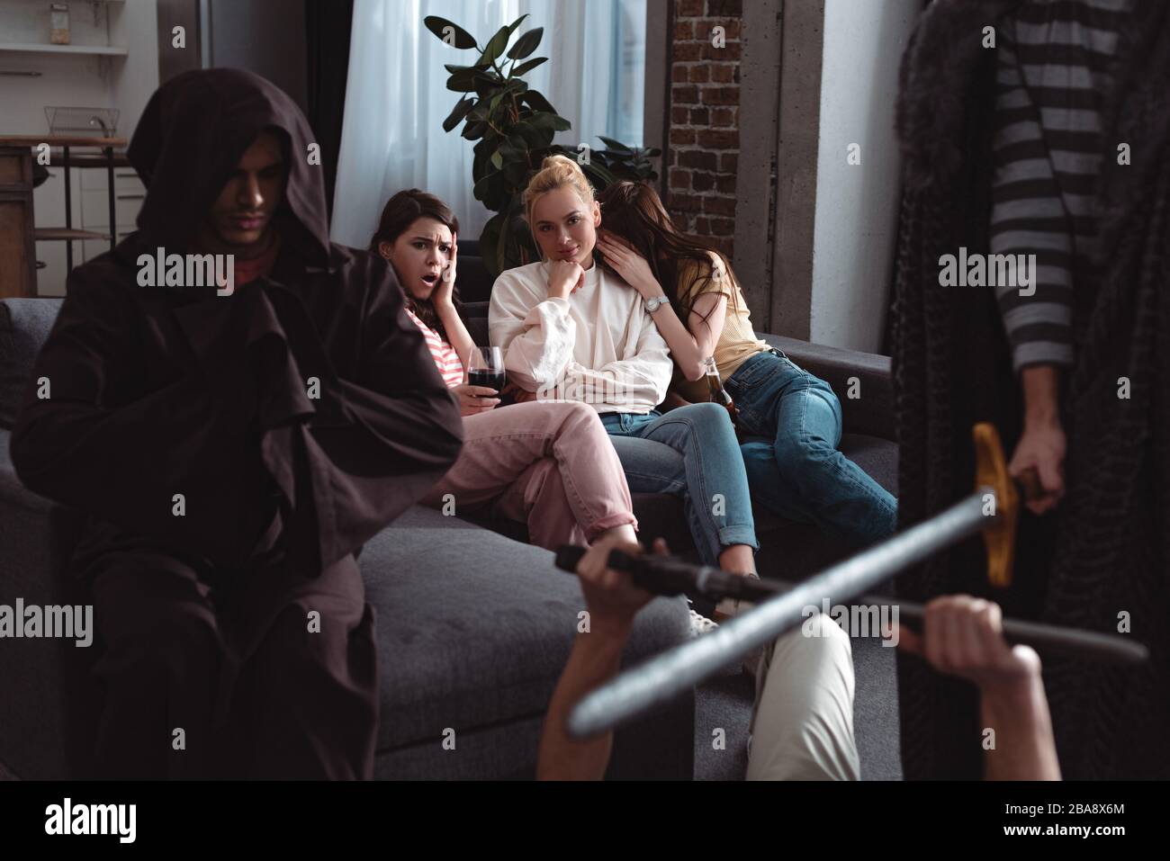 cropped view of men fighting with toy swords near guy in costume of monk while worried girls sitting on sofa Stock Photo