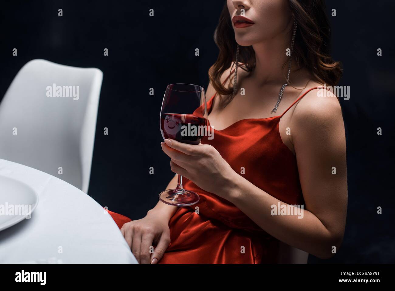 Cropped view of elegant woman in red dress holding wine glass at table  isolated on black Stock Photo - Alamy