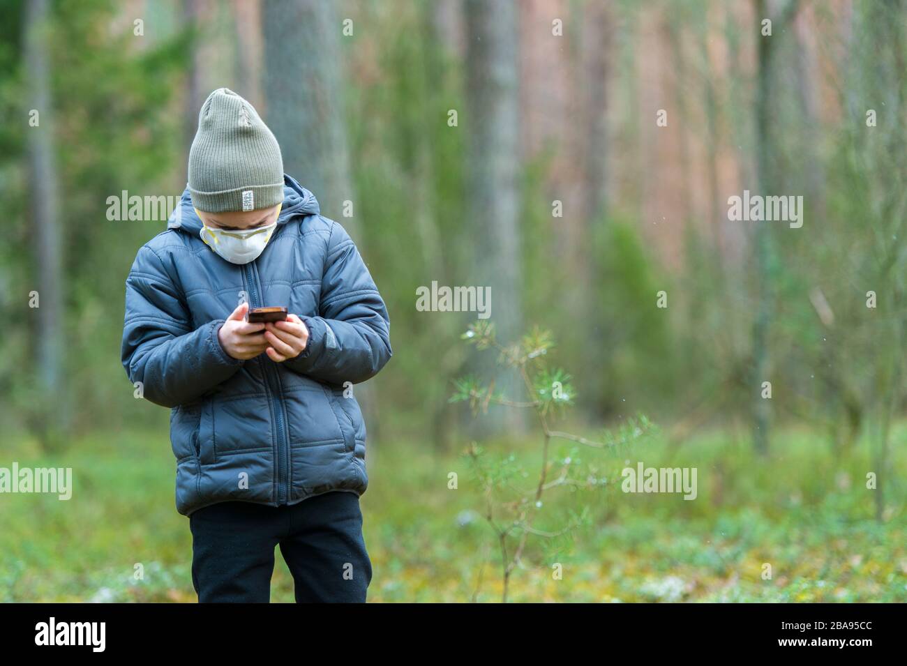 Kid with mask in their mouths due to coronavirus disease and play mobile games on a black phone in a forest Stock Photo
