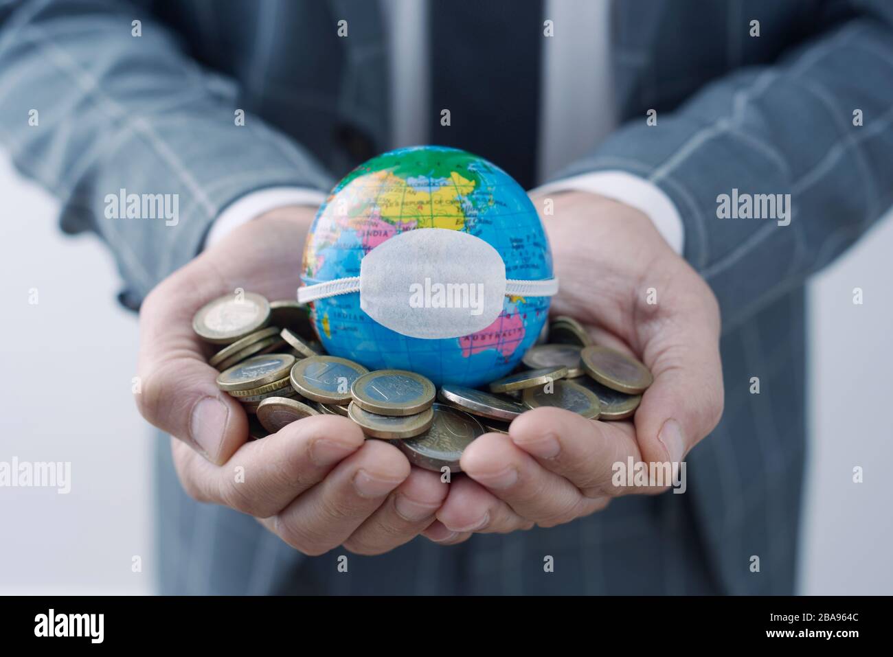 businessman holding a world globe with a protective mask on top of a handful of euro coins, depicting the economic consequences of the coronavirus pan Stock Photo
