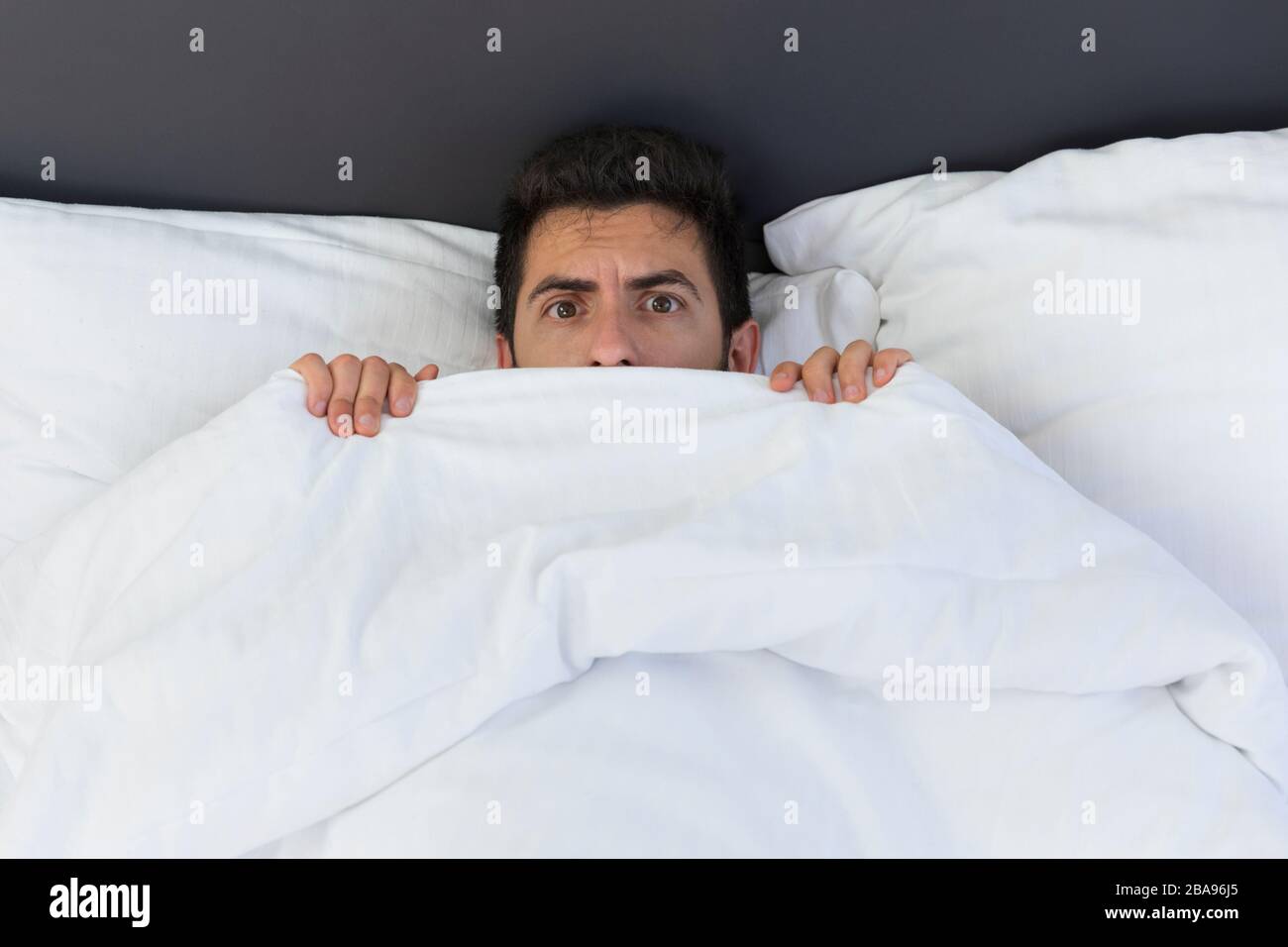 A young man is hiding in bed under a blanket at home. Fear and surprise