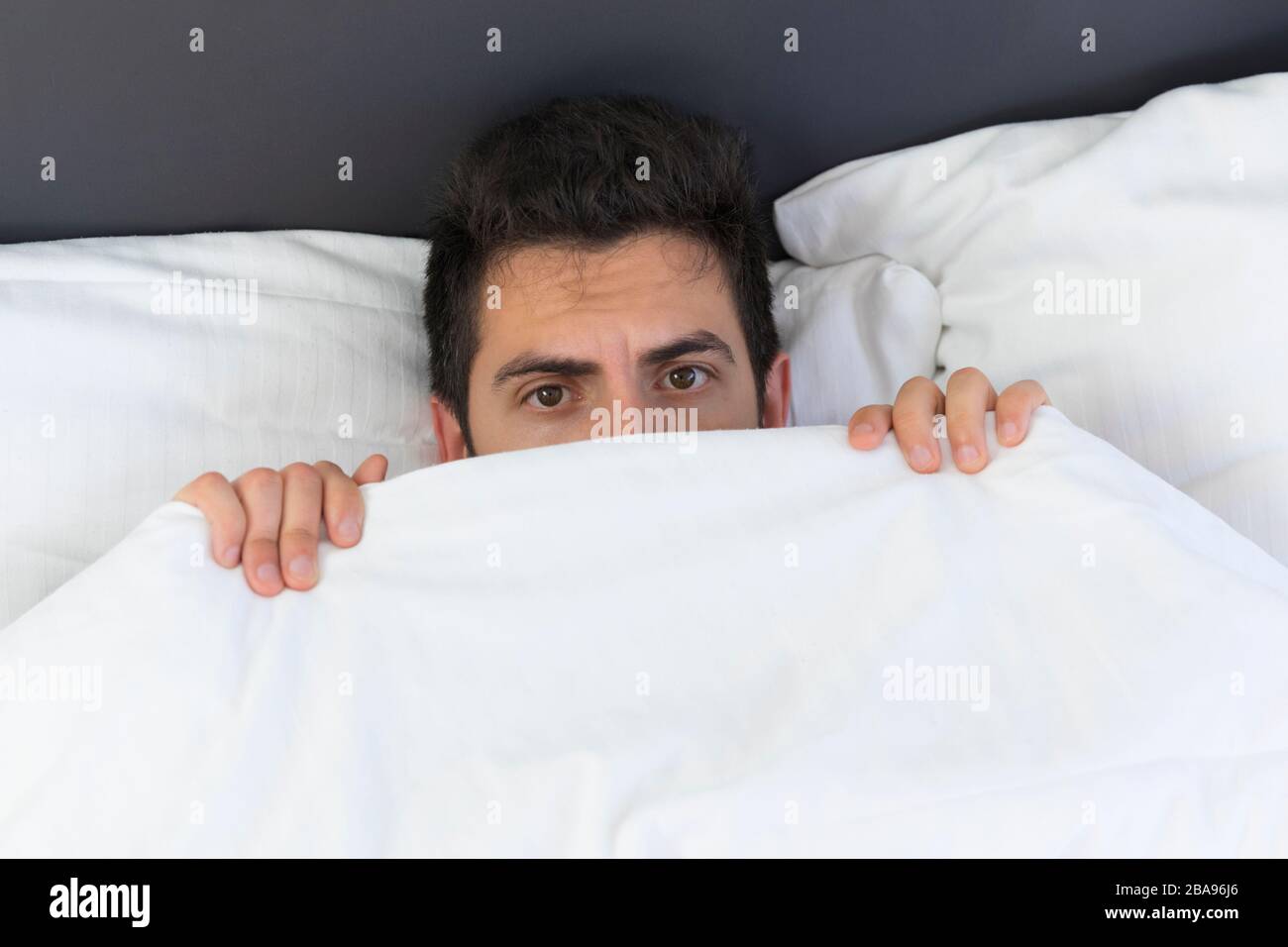 A young man is hiding in bed under a blanket at home. Fear and surprise are visible in his eyes. Stock Photo