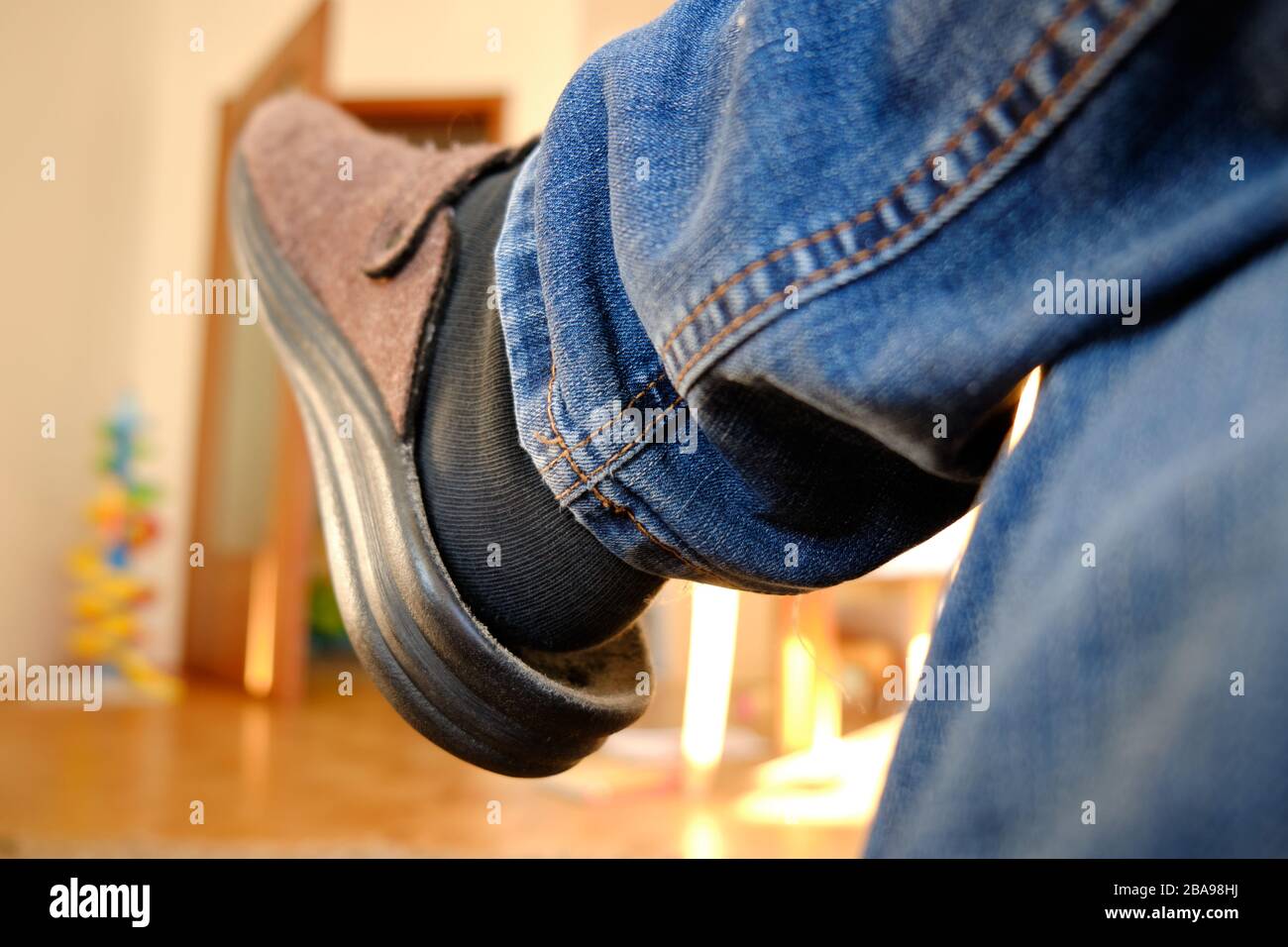 Closeup of right leg of a man wearing blue jeans and slippers while sitting bored inside the apartment on a sunny day during corona crisis. Seen in Ge Stock Photo