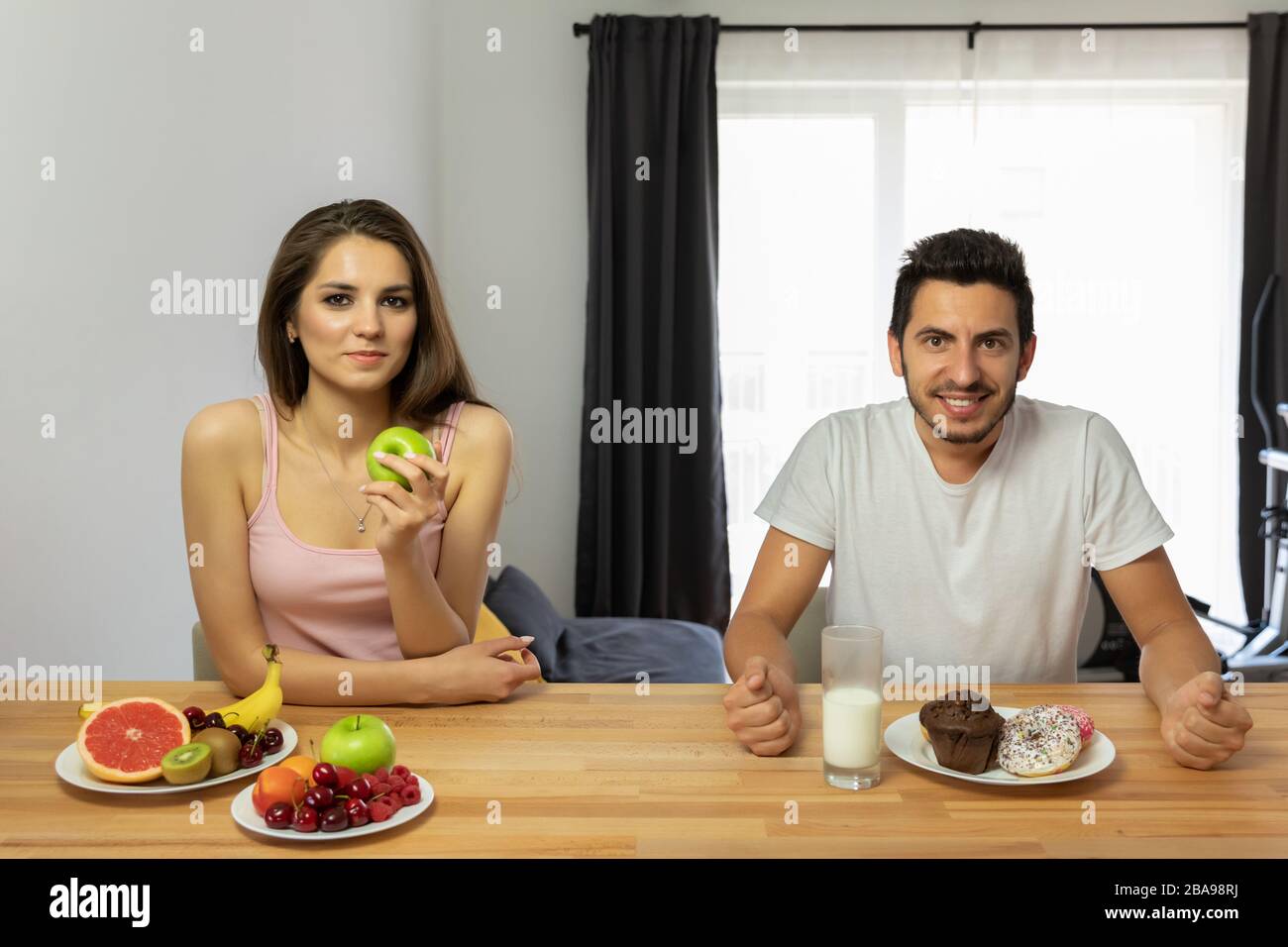 A man leads a wrong lifestyle, he eats donuts with icing for breakfast. The girl prefers fresh fruits and berries, she is a vegan. The confrontation o Stock Photo
