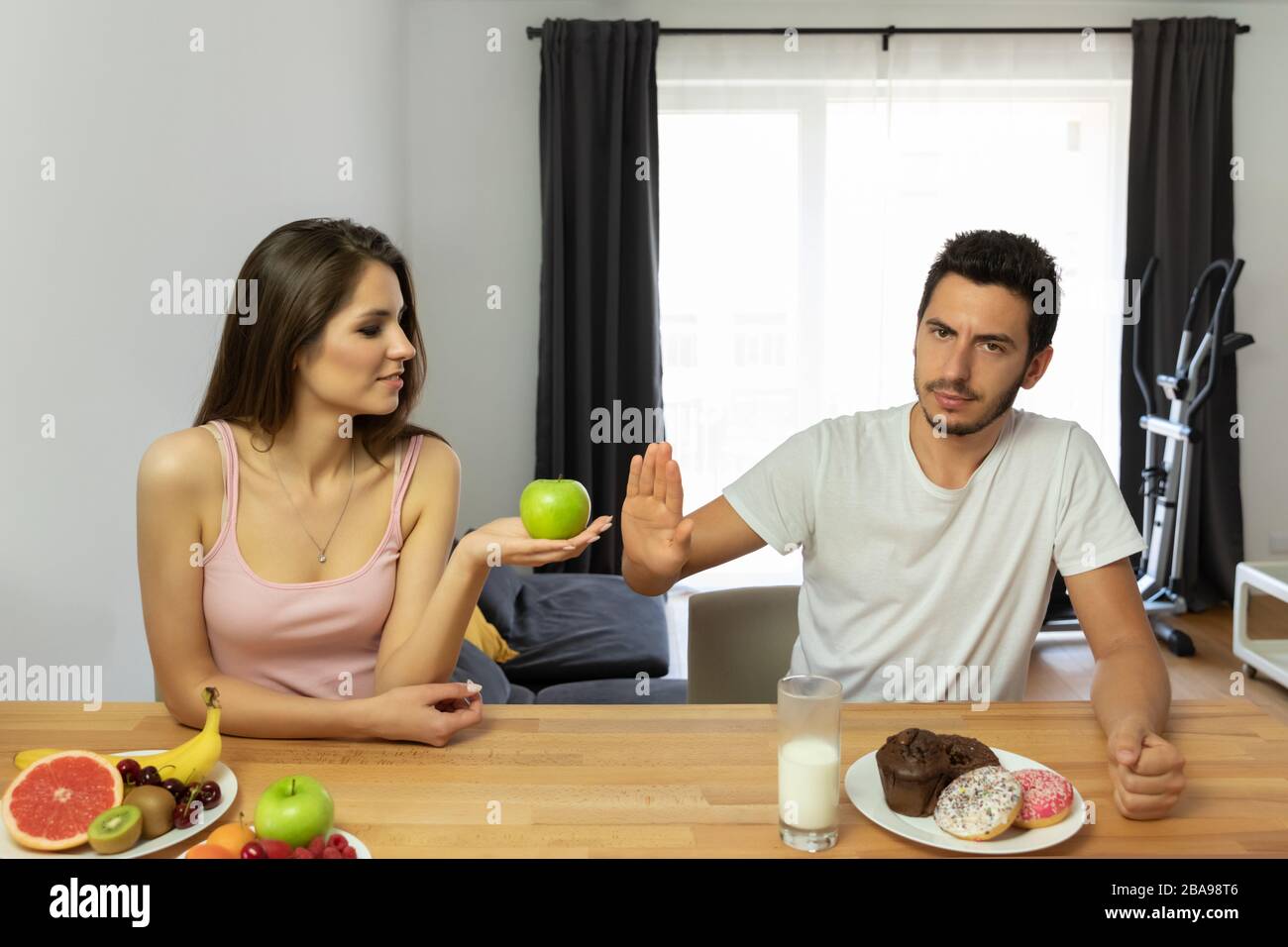A man leads a wrong lifestyle, he eats donuts with icing for breakfast. The girl offers him to try fresh fruits, but he refuses. The confrontation of Stock Photo
