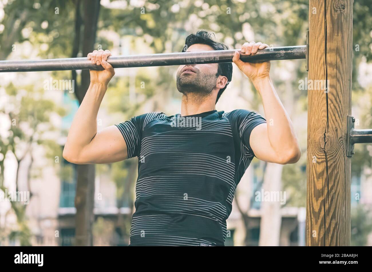 Young man making pull-up strength training exercise - fitness guy working out his arm muscles on outdoor park gym doing chin-ups / pull-ups CrossFit Stock Photo