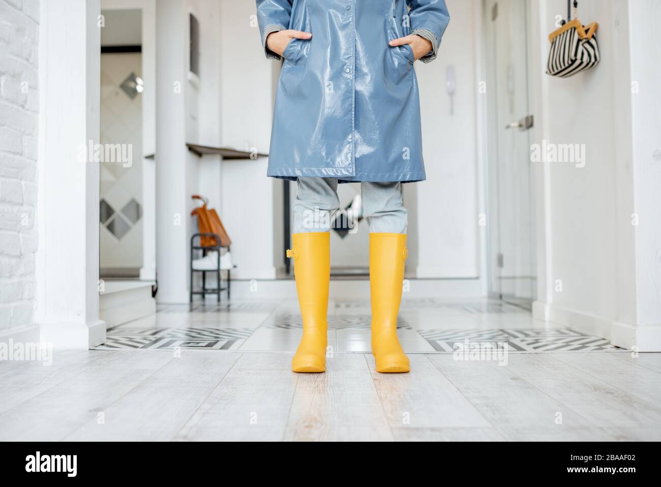 Woman in yellow rubber boots and raincoat standing in the corridor at home, ready to walk outside in rainy weather Stock Photo