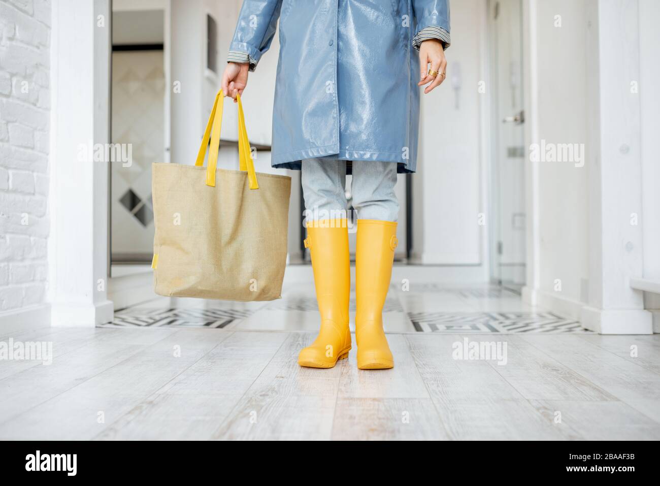 Woman in yellow rubber boots and raincoat standing with shopping bag in the corridor at home, ready to walk outside in rainy weather Stock Photo