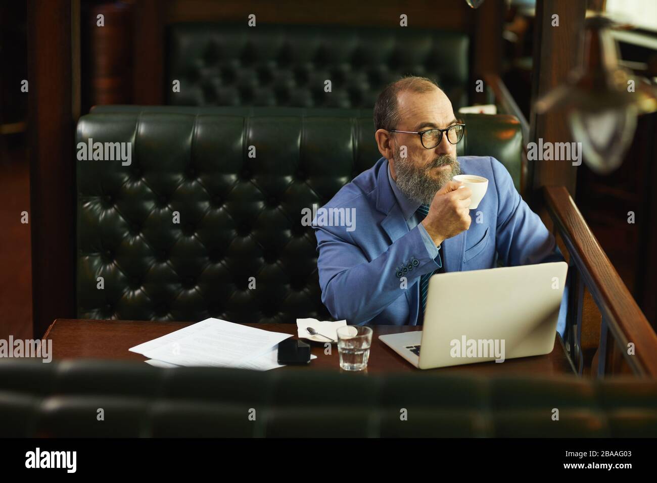 Senior bearded businessman drinking coffee and thinking about something while working with laptop in cafe Stock Photo