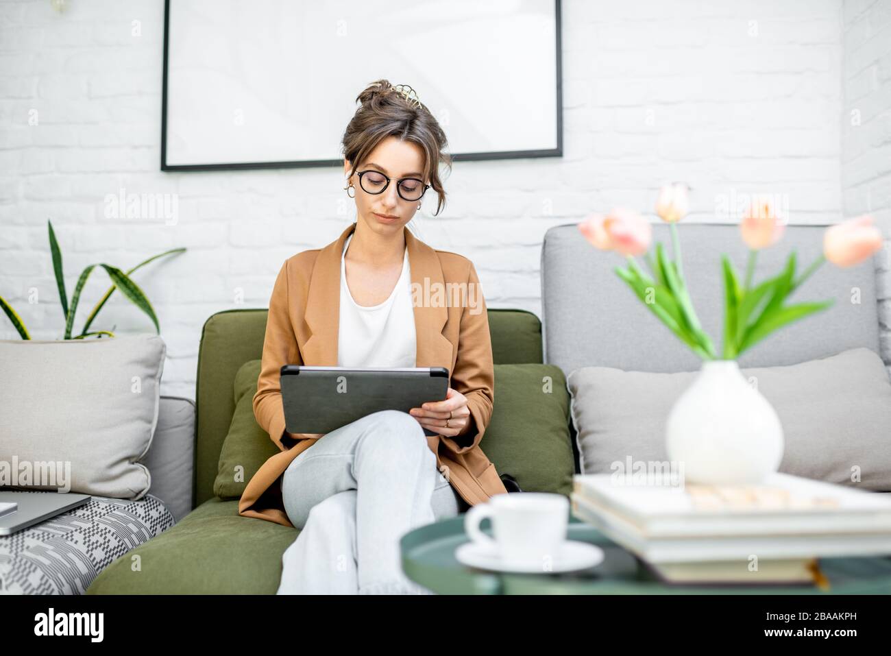 Young creative and well-dressed woman working on a digital tablet while sitting on the comfortable couch at home. Concept of a freelance and creative work online Stock Photo