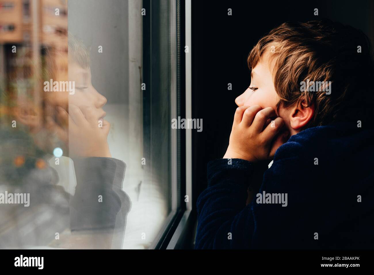 Sad and melancholic boy looks through a window in his apartment during ...