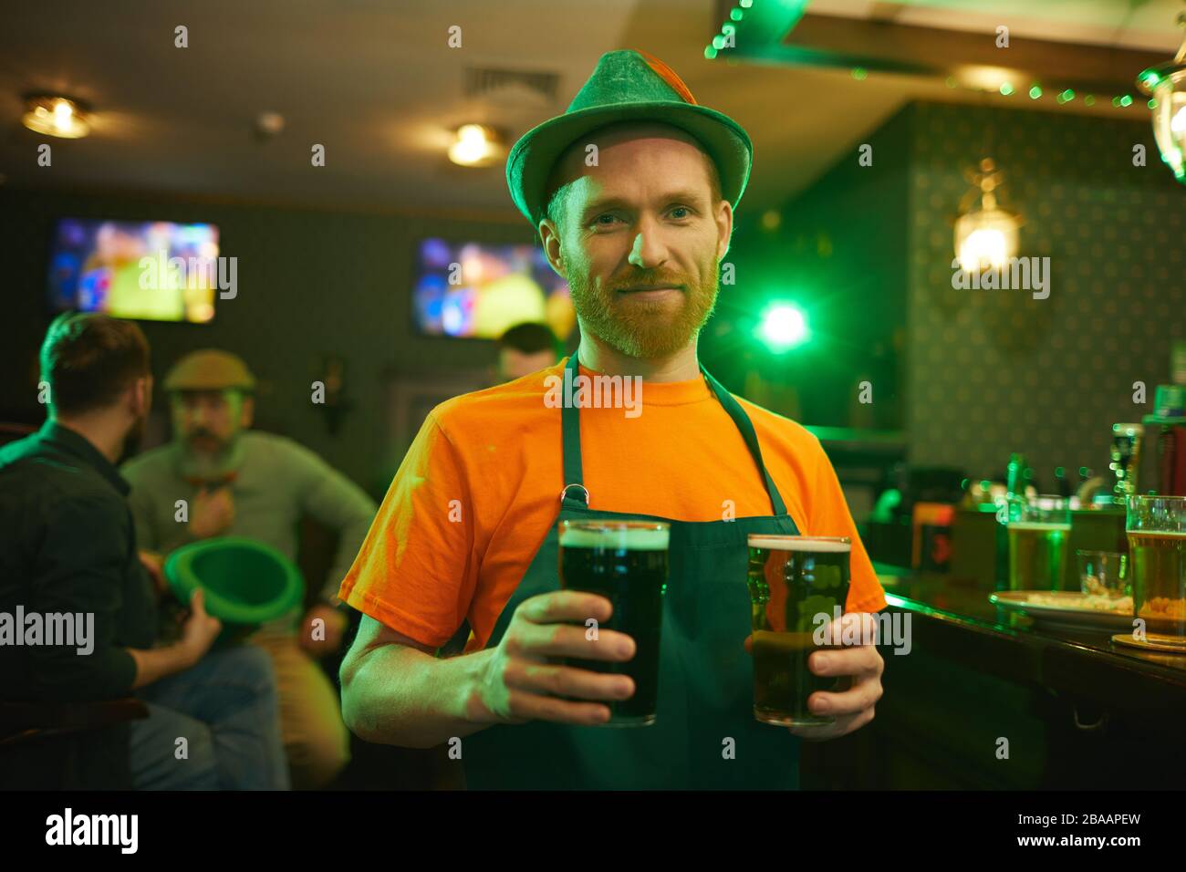 Portrait of young bearded waiter in green apron and hat holding two glasses of beer and smiling at camera while working in the pub Stock Photo